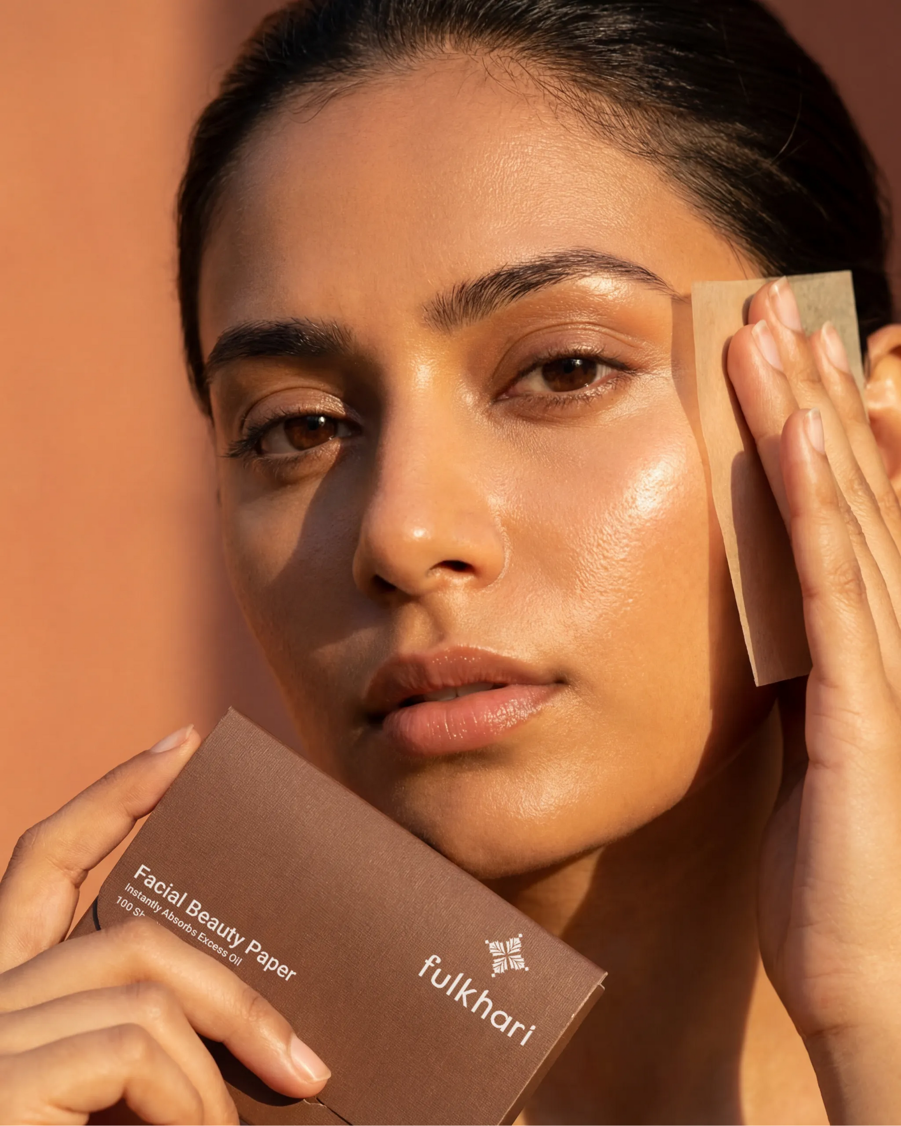 Model with glowing skin using a Fulkhari oil blotting sheet to instantly absorb excess sebum and mattify the T-zone without disturbing makeup.