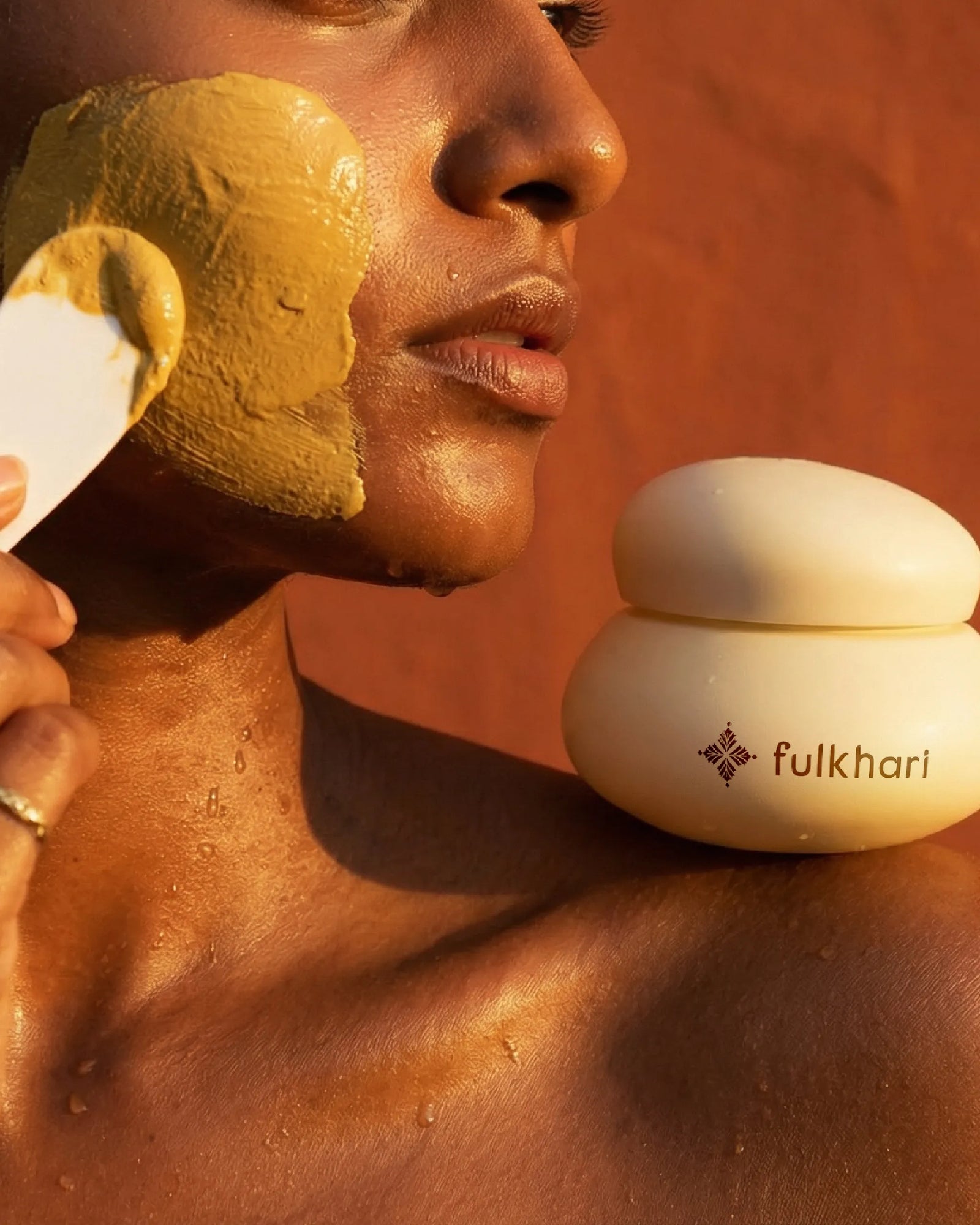 Model with glowing skin applying the yellow turmeric and Multani Mitti clay mask to her cheek for dark spots.